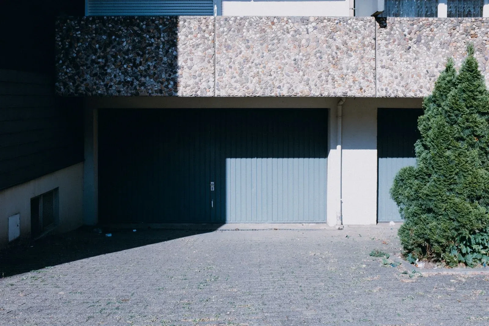 Two gray-blue vertical-plank garage doors under a pebble-textured concrete overhang, half in shadow, beside a green shrub and a paved lot. — Orono