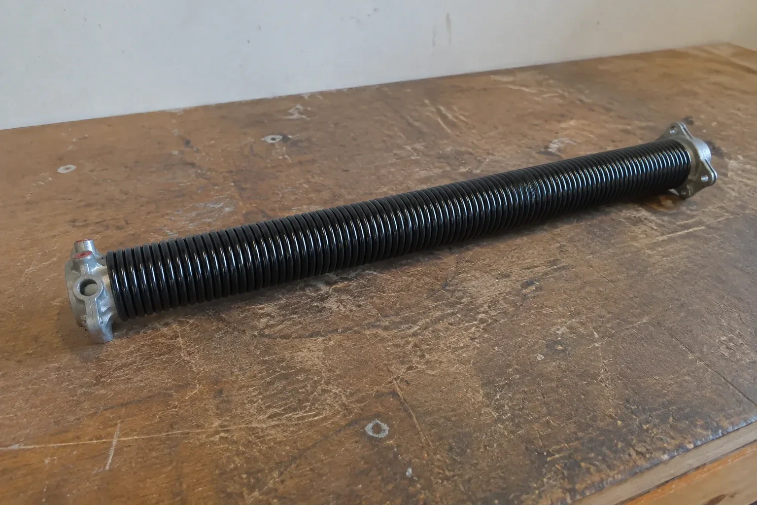 Detached black coil spring with metal end fittings on a workbench. — Torsion Spring Replacement, Clarington