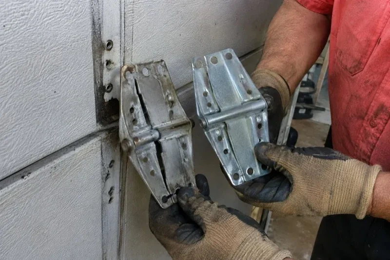 Gloved hands holding two metal garage-door hinges side by side, one discolored with a visible crack and the other bright silver. — Garage Door Hinge Replacement, Clarington